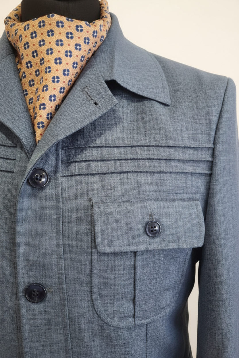 Gros plan veste safari bleu ardoise micro pied-de-poule trois tucks horizontaux décoratifs en relief poche poitrine bouton marine ascot géométrique multicolore or orange marine vert revers lapel fond blanc pur studio détail artisanat luxe