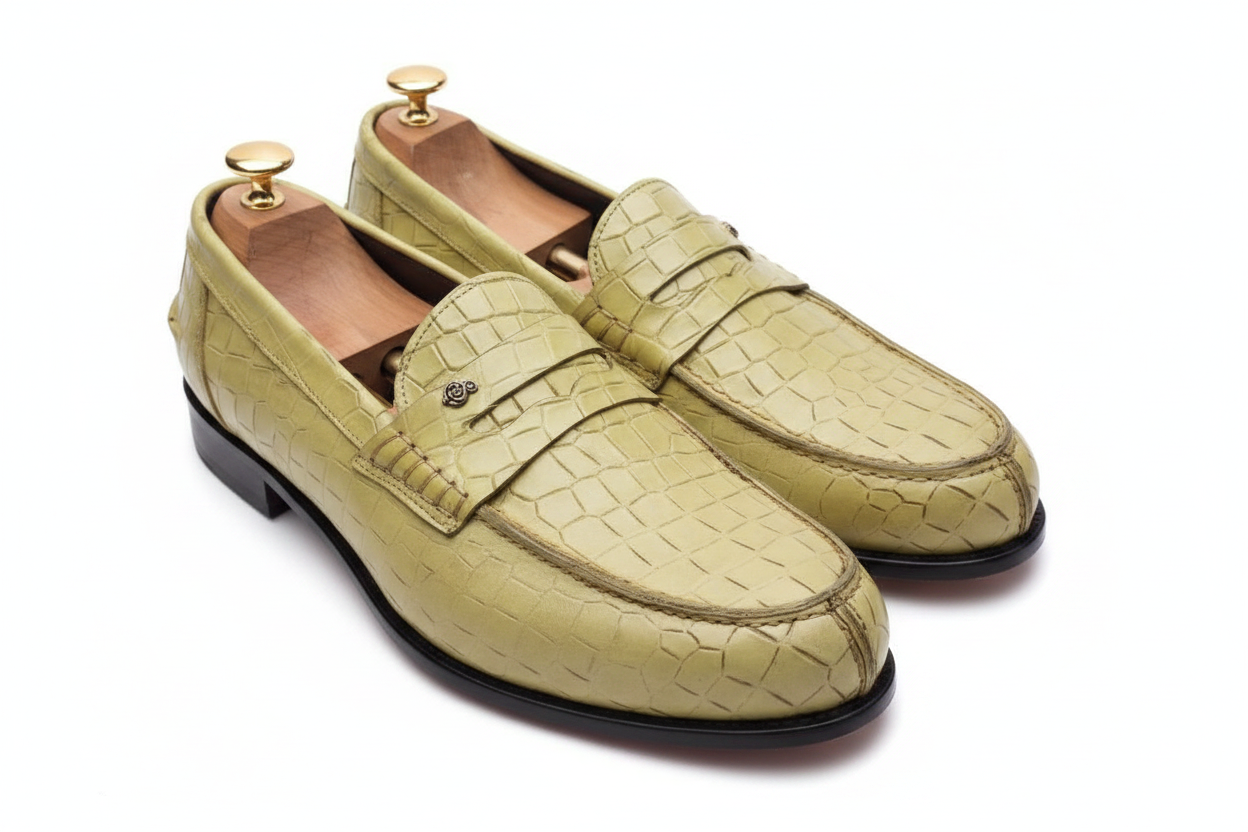 Mocassins penny loafer cuir croco embossé vert olive chartreuse vue plongeante légèrement élevée écailles croco vamp visible strap penny médaillon logo rose doré embauchoirs cèdre bouton doré intérieur caramel fond blanc pur studio luxe
