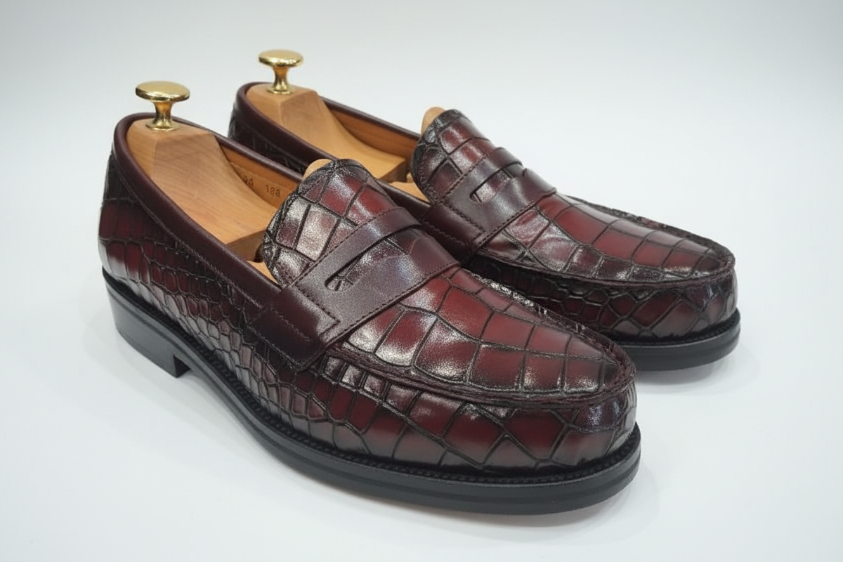 Mocassins penny loafer cuir crocodile bordeaux oxblood cognac patine satin vue trois-quarts avant gauche légèrement surélevée deux zones écailles vamp grandes rectangulaires grille damier tridimensionnelles côtés organiques irrégulières plus petites transition beauté patine satin cognac ambre reflets lumineux sillons sombres strap chocolat barre large sans fente couture mocassin welt chocolat semelle noire épaisse talon empilé embauchoir cèdre bouton doré lustré fond blanc pur studio luxe paysage