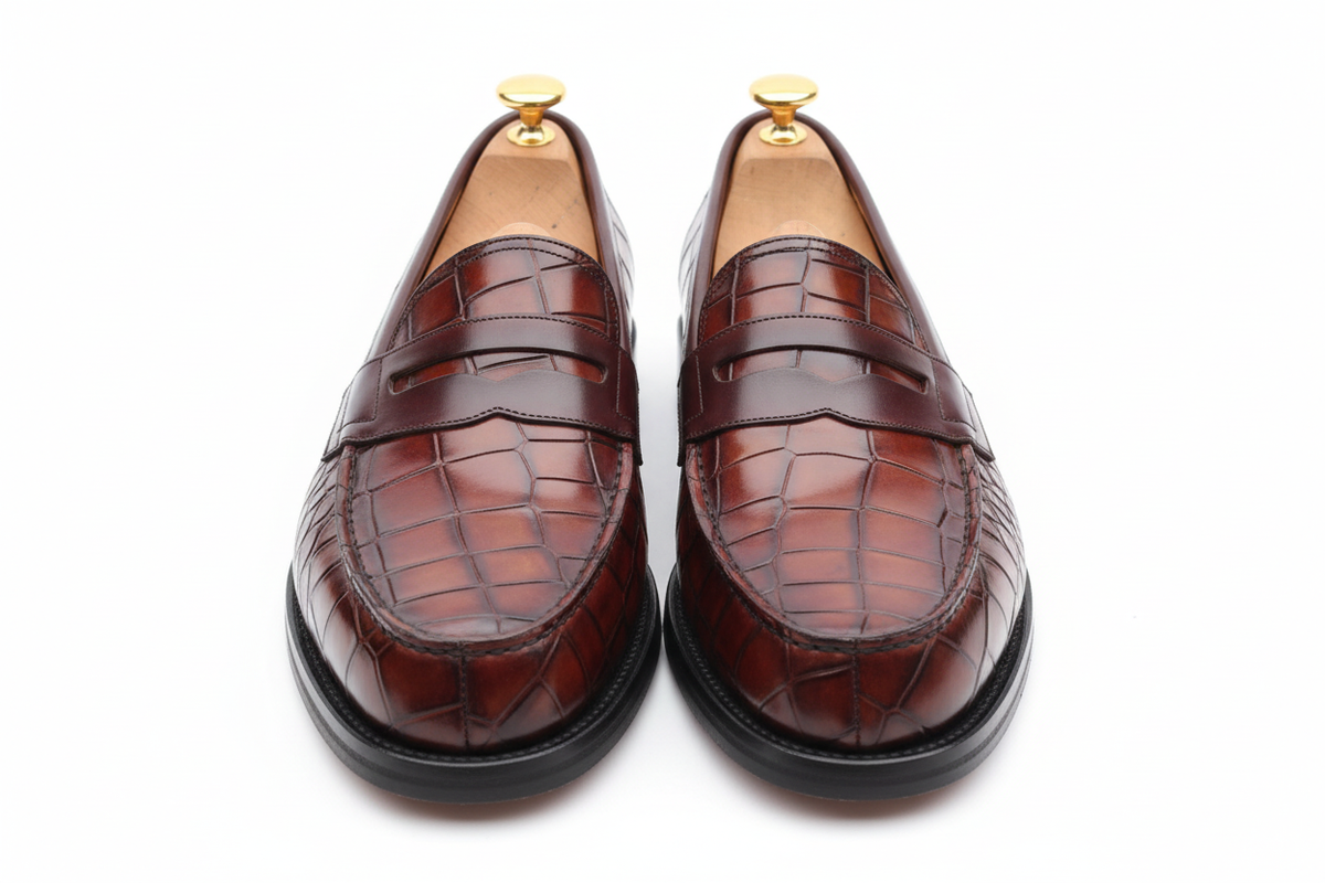 Mocassins penny loafer cuir crocodile bordeaux oxblood cognac patine vue frontale symétrique bout carré rectangulaire très large grandes écailles damier grille tridimensionnelles vamp large double barre strap chocolat lisse welt piping sombre semelle plate noire épaisse large embauchoirs cèdre bouton doré unique grand plat intérieur tan fond blanc pur studio luxe paysage