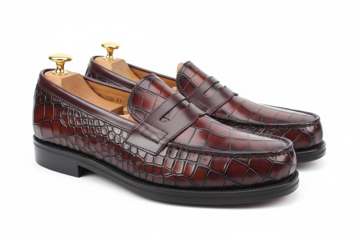 Mocassins penny loafer cuir crocodile bordeaux oxblood cognac patine vue large basse angle profil flanc gauche deux chaussures côte à côte silhouette complète grandes écailles irrégulières rectangulaires carrées côtés profil organiques strap chocolat lisse barre large couture mocassin welt chocolat noir piping semelle noire épaisse talon empilé bas large embauchoir cèdre bouton doré unique grand brillant fond blanc brillant pur studio luxe paysage