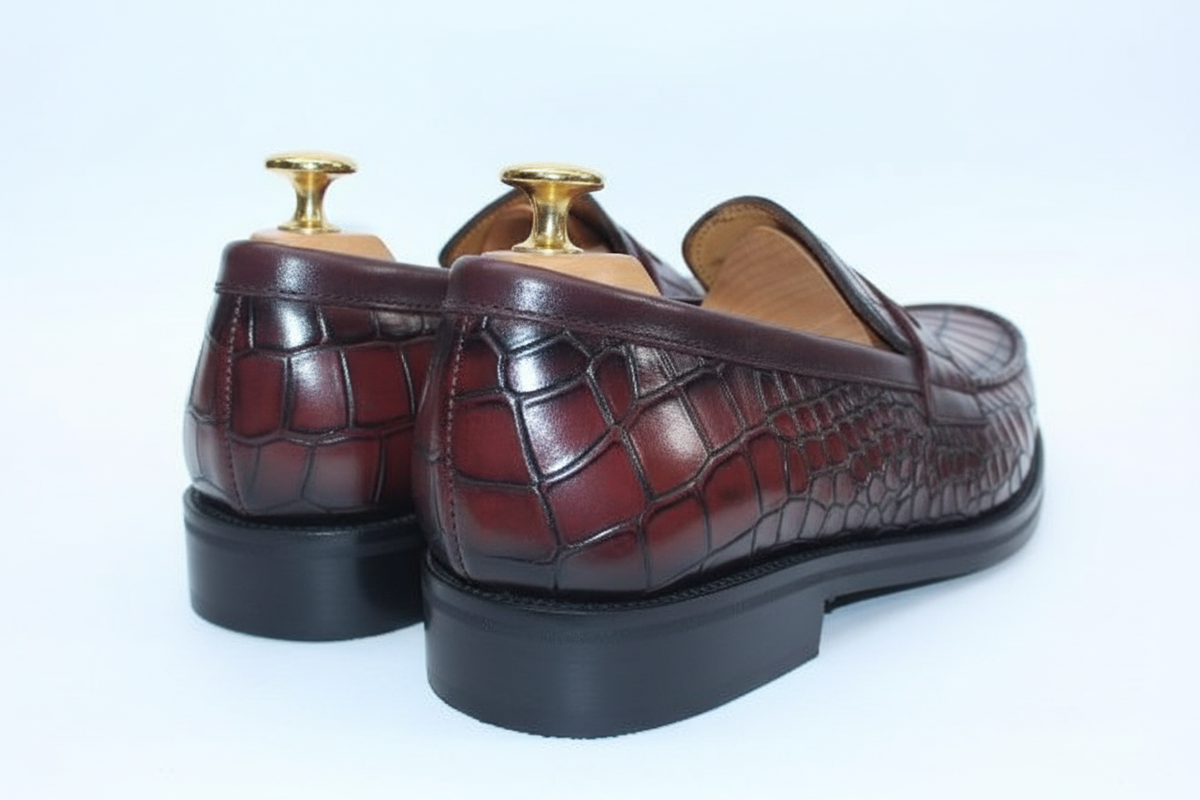 Mocassins penny loafer cuir crocodile bordeaux oxblood rouge cramoisi creux écailles cognac ambre crêtes patine dégradé vue arrière gauche très basse angle sol contrefort cuir laqué brillant haute gloss miroir sombre chocolat contraste texture crocodile talon empilé épais large noir semelle plate épaisse noire welt chocolat embauchoirs cèdre bouton doré unique grand brillant intérieur tan peach fond blanc brillant pur studio luxe paysage