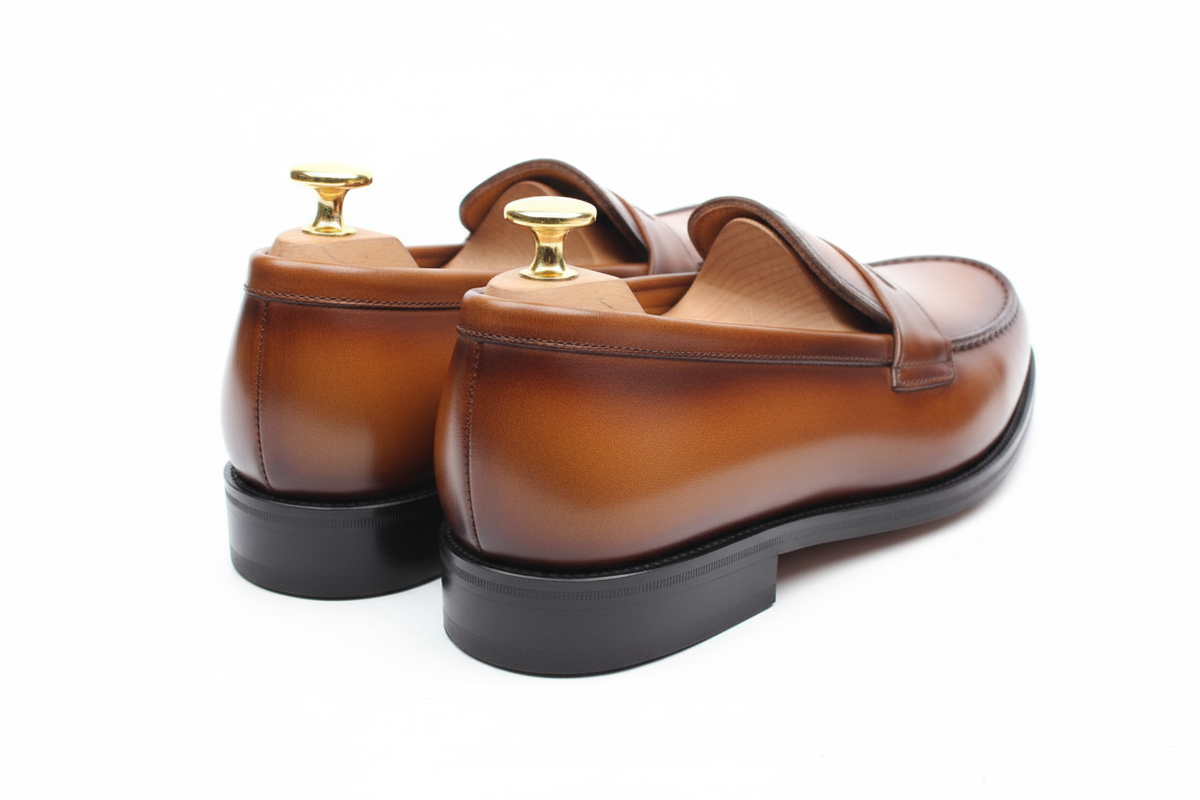 Mocassins penny loafer cuir lisse grain naturel tan cognac ambre patine main dégradé bords noircis artisanal vue arrière trois-quarts contreforts barrel arrondi lisse tan bords noircis gradient quartiers arrière grain naturel légèrement pebbled embauchoirs cèdre deux boutons dorés grand et petit symétriques brillants intérieur tan talon empilé épais large noir plat welt Goodyear burnished sombre fond blanc pur studio luxe paysage
