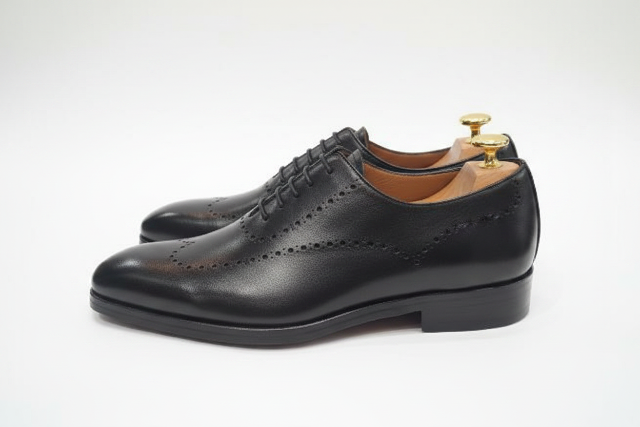 Oxford brogue noir vue profil latéral côté gauche deux chaussures décalées semelle cuir welt visible embauchoir cèdre deux boutons dorés laiton perforations brogue côté fond blanc pur studio luxe